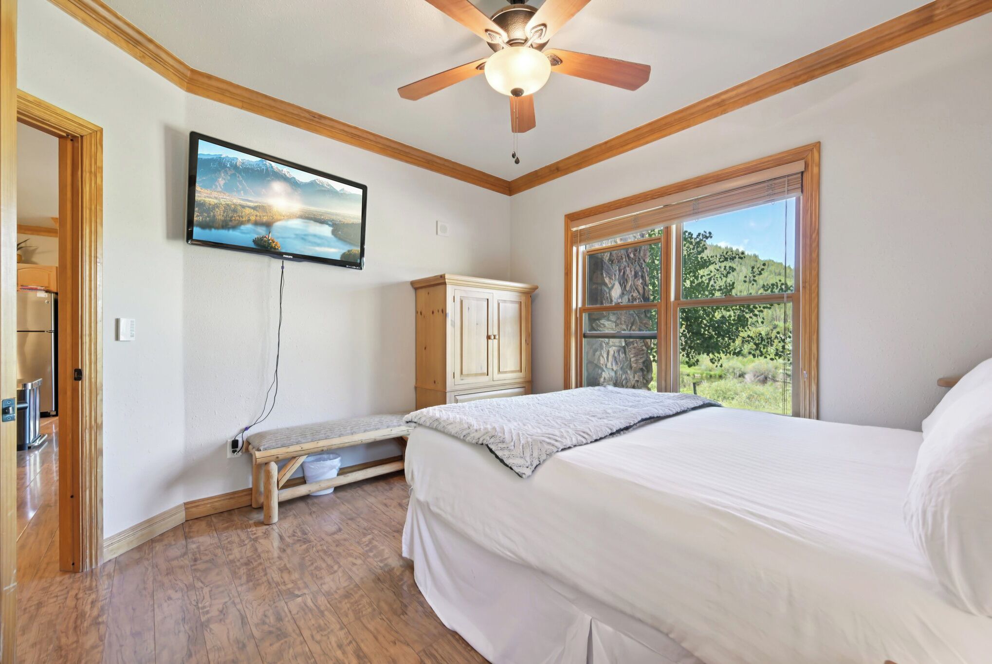 Ein Schlafzimmer mit einem Bett, einem Deckenventilator, einem an der Wand montierten Fernseher, einem Fenster mit Blick auf Grünflächen und einer hölzernen Bank.