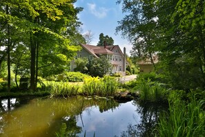 Une grande maison avec un étang au premier plan, entourée d’une végétation luxuriante et d’arbres.