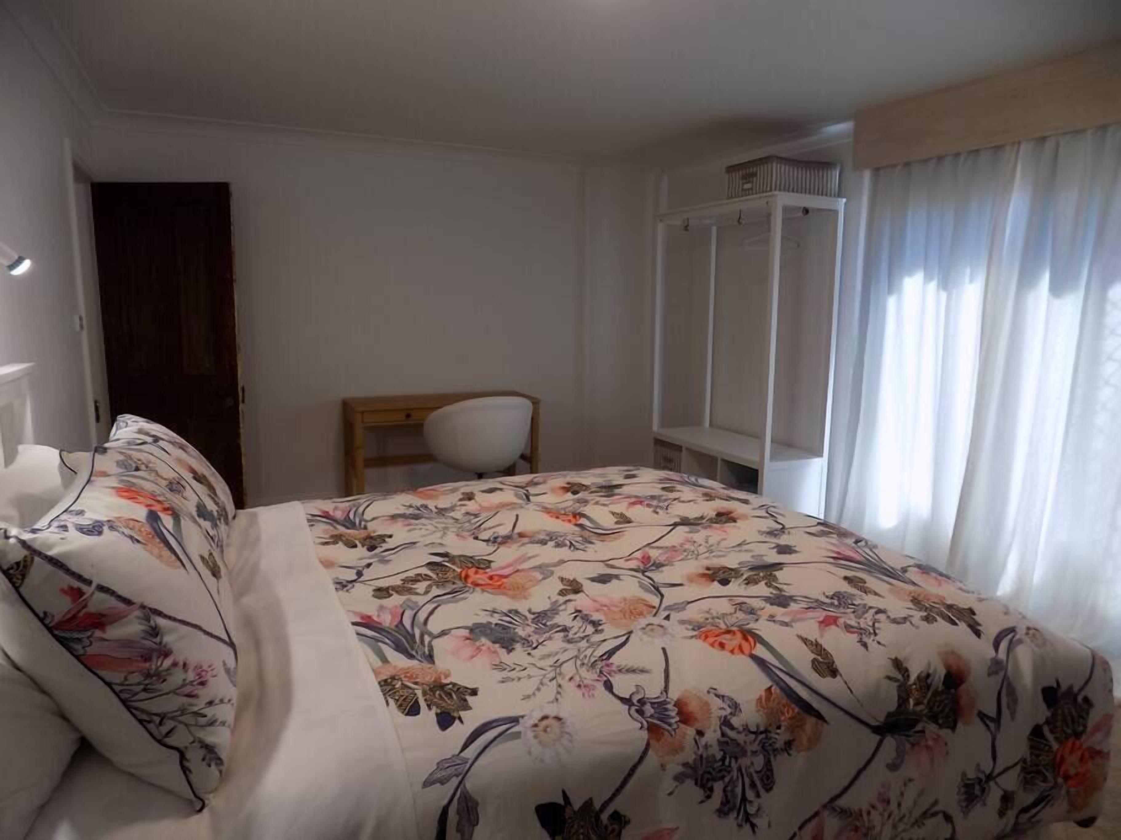Ein Schlafzimmer mit floraler Bettdecke, einem Holzsessel, einem Kleiderschrank und einem Fenster mit Vorhängen.