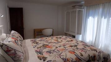 A bedroom with a floral bedspread, a wooden chair, a wardrobe, and a window with curtains.
