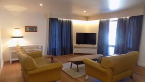 Un salon avec deux fauteuils jaunes, une table basse, un téléviseur à écran plat posé sur un meuble blanc et une lampe sur une console blanche.