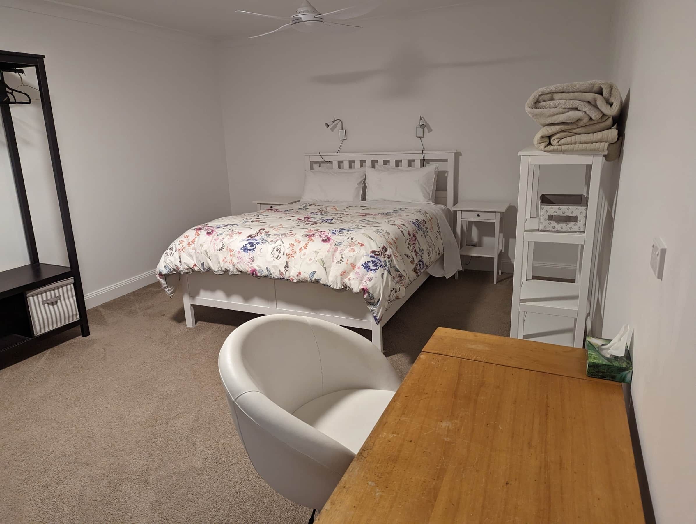 A bedroom with a bed, a chair, a wooden table, a white shelf, and a ceiling fan.