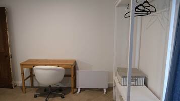 A room with a wooden desk, a white office chair, a white radiator, and a white shelving unit with a hanging clothes rack.