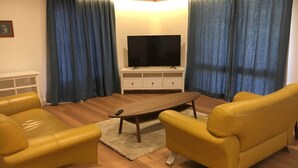 Ein Wohnzimmer mit Fernseher auf einem weißen Schrank, gelben Sofas, einem Holztisch und blauen Vorhängen.