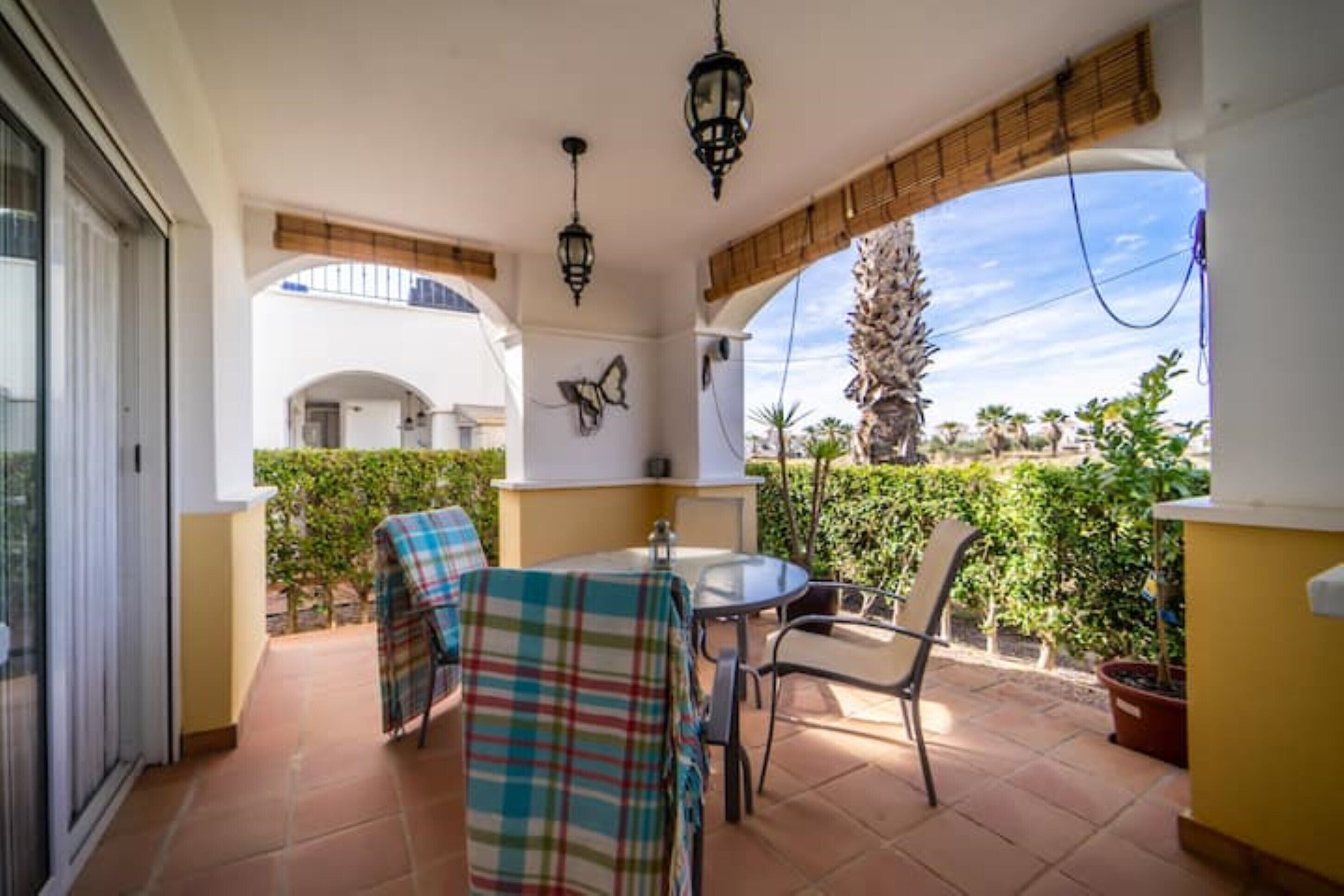Un balcón con mesa y sillas, un adorno de pared con forma de mariposa y vistas a una palmera y follaje.