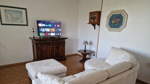 Una sala de estar con un sofá blanco, un mueble de madera, un televisor mostrando una aplicación de streaming y una mesita con una lámpara.
