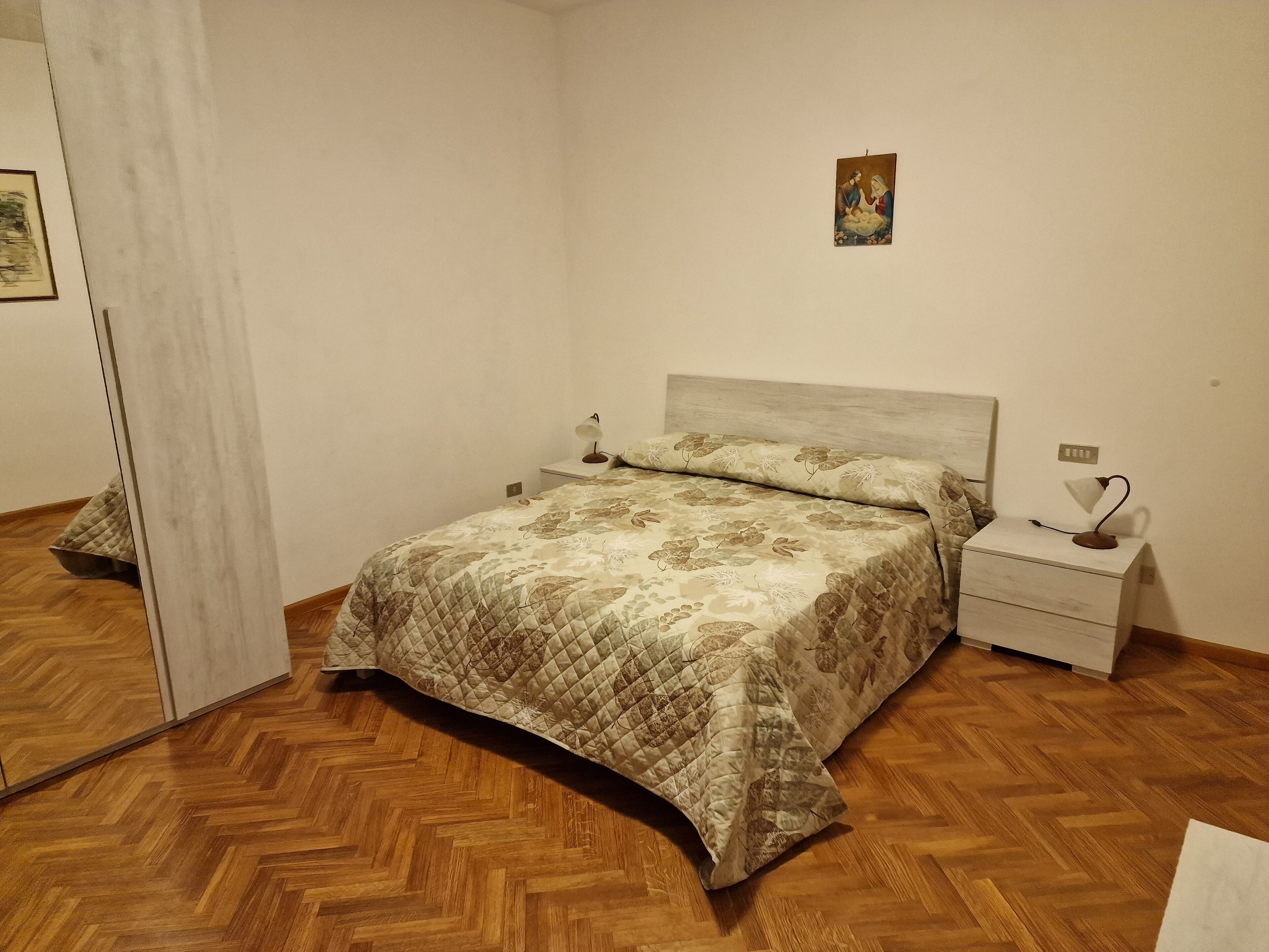 Une chambre avec un parquet en bois disposé en motif chevrons, un lit recouvert d’une courtepointe matelassée, une table de chevet avec une lampe et un petit tableau accroché au mur.
