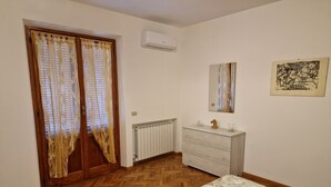 Un dormitorio con suelo de parqué, una cama con edredón estampado con motivos vegetales, un aparador blanco, un radiador y una ventana con cortinas de encaje.
