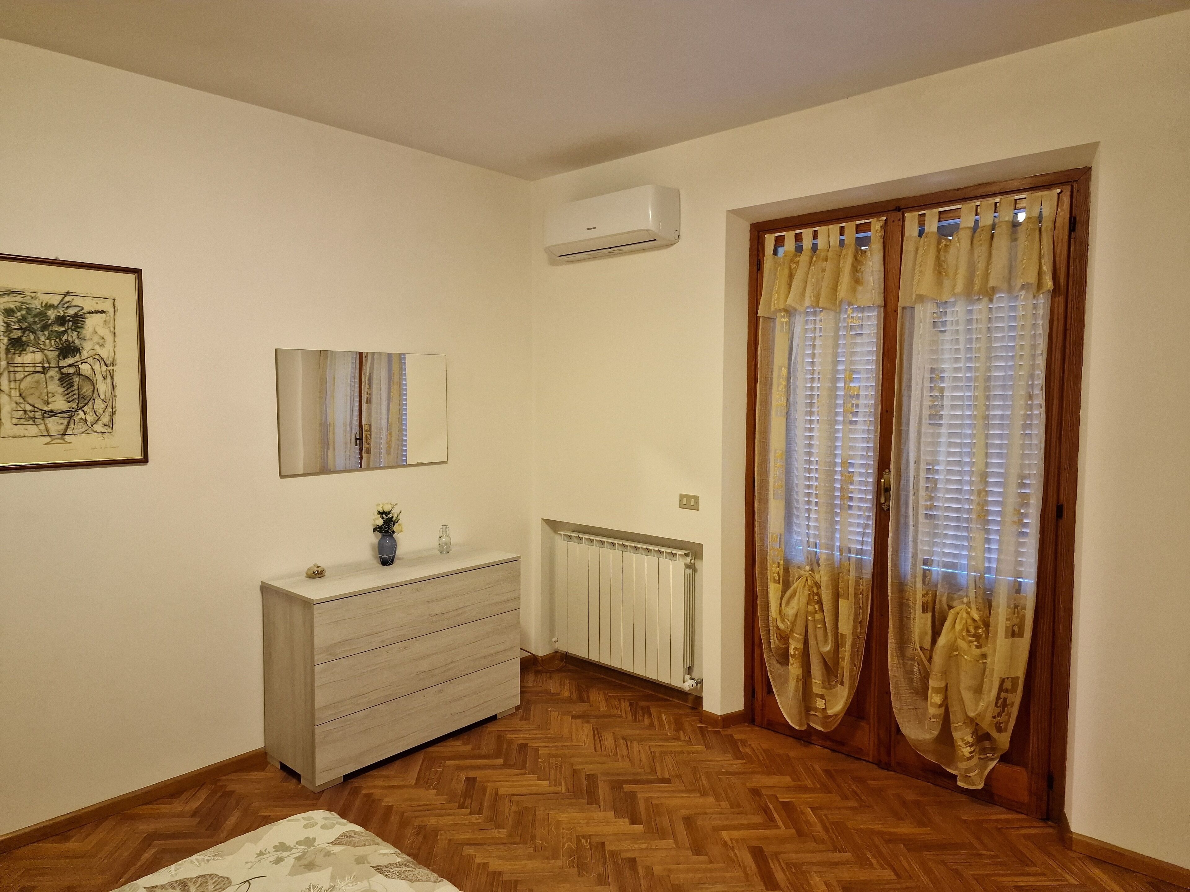 A room with a wooden herringbone floor, a white dresser with a vase of flowers, a framed abstract painting, a mirror, and a radiator.