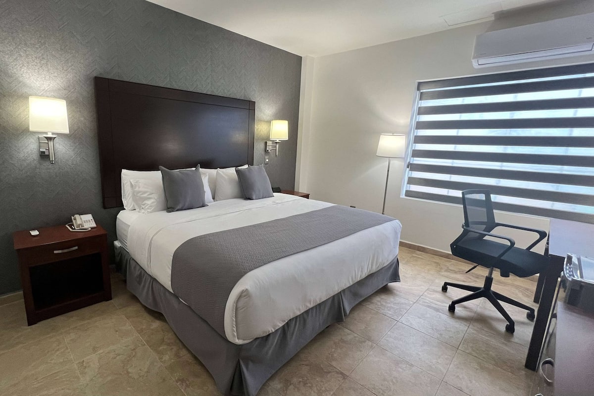 Habitación de hotel con una cama grande, dos mesitas de noche con lámparas, un escritorio con una silla y una ventana con persianas.