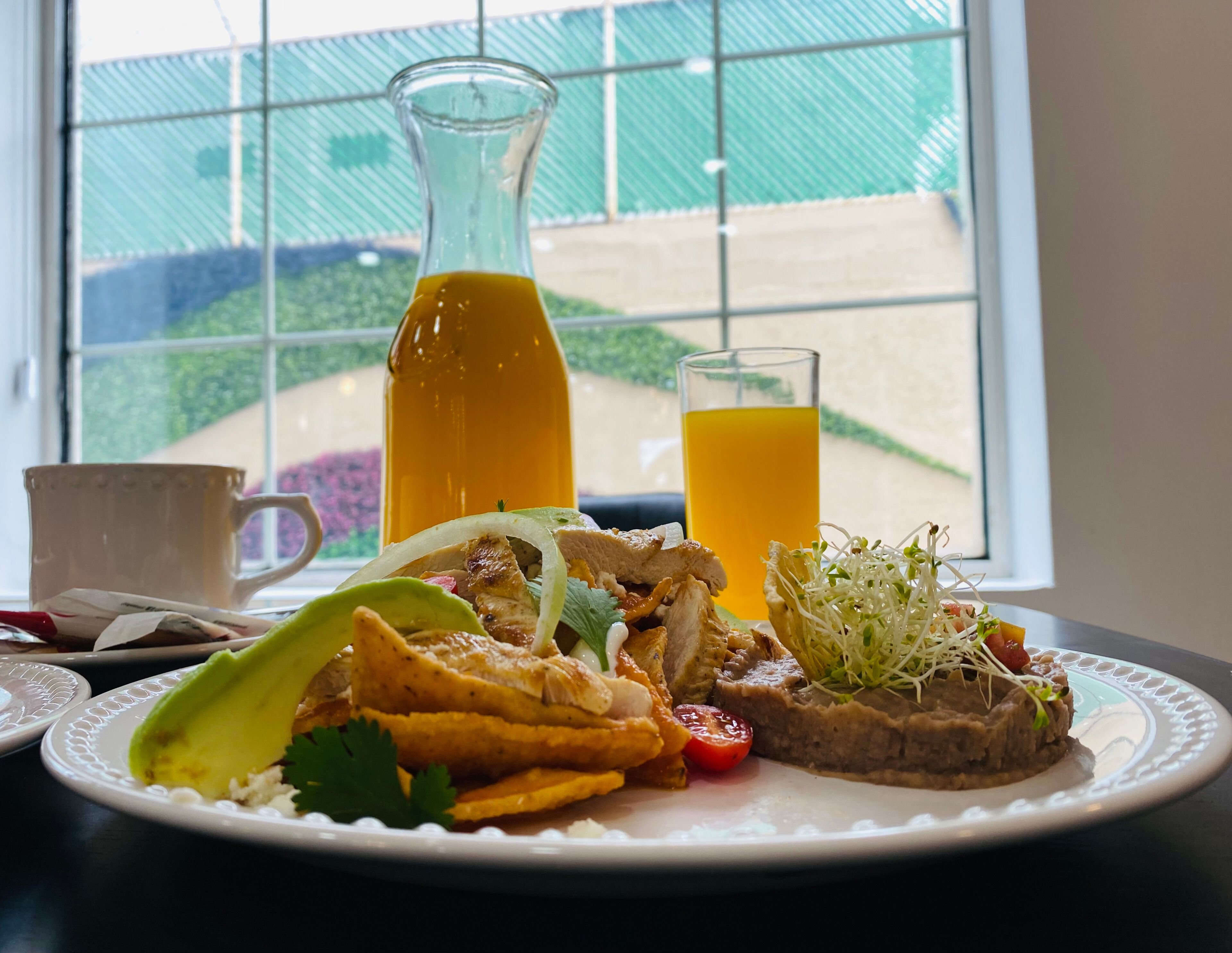 A breakfast plate with a glass of orange juice, a bottle of orange juice, and a cup on a table by a window with a view of a garden.