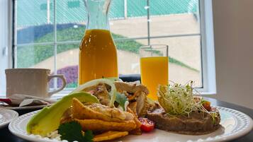 A breakfast plate with a glass of orange juice, a bottle of orange juice, and a cup on a table by a window with a view of a garden.