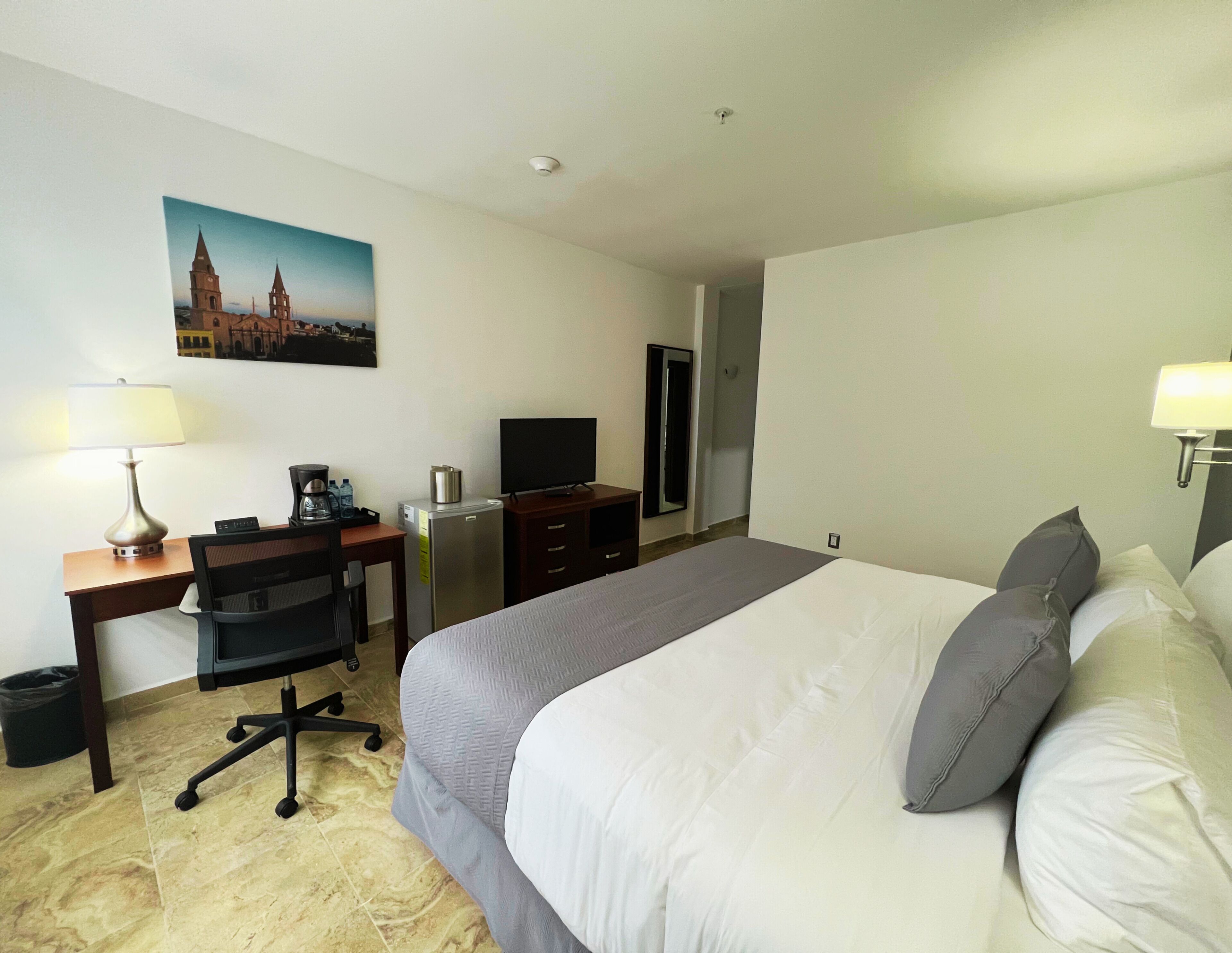 Une chambre d’hôtel avec un lit, un bureau, une chaise, une télévision et un tableau représentant un bâtiment accroché au mur.