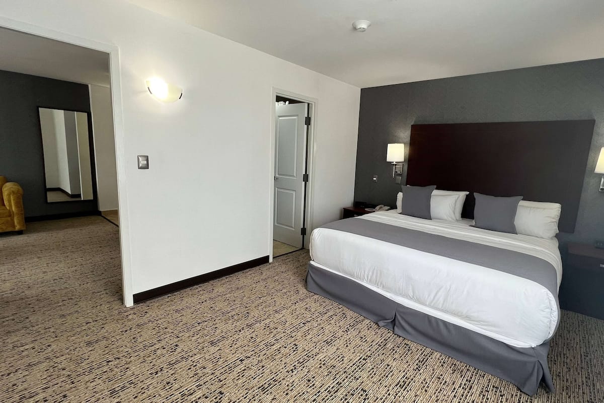 Habitación de hotel con una cama grande, una mesita de noche, una lámpara y una puerta que da a otra habitación.