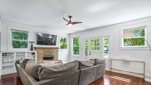 A living room with a stone fireplace, a grey sofa, wooden flooring, and large windows with greenery outside.