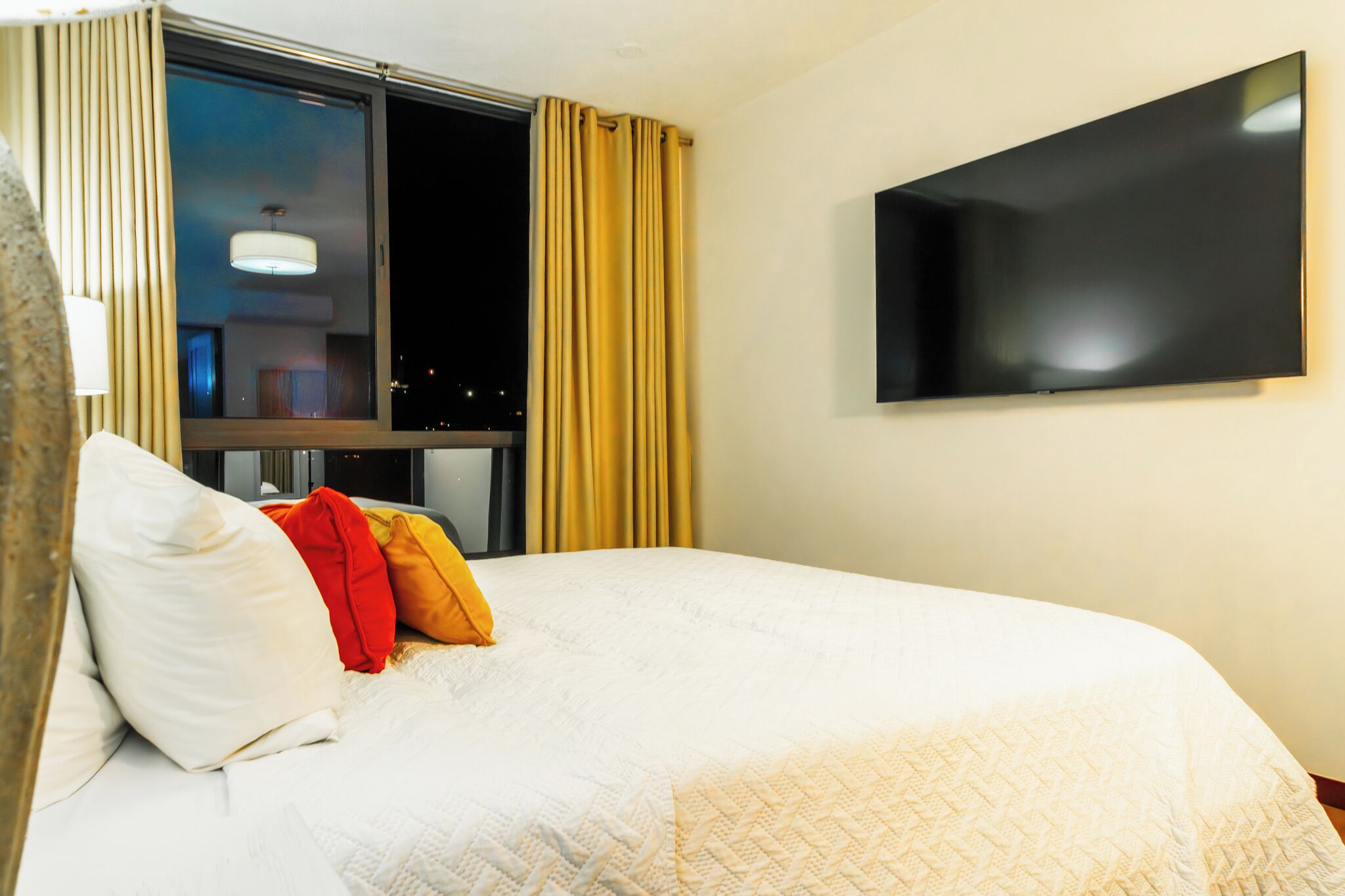 Habitación de hotel con cama, televisor montado en la pared, ventana con cortinas y vista al cielo nocturno.