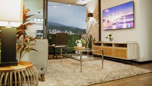 A modern living room with a glass coffee table, a sofa, a TV mounted on the wall, and a view of a cityscape through a glass door. - Altos Tower Apartamento de 3 habitaciones (San Salvador)