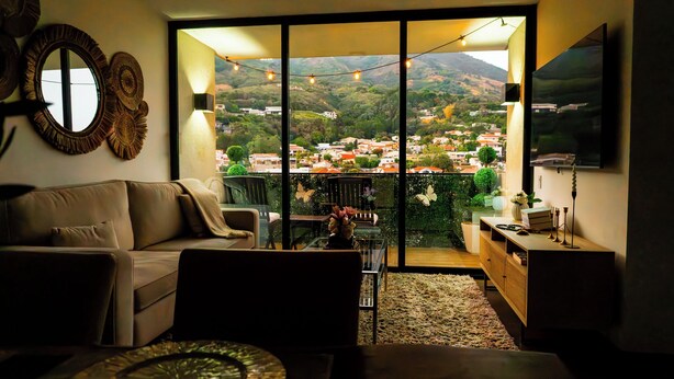 A living room with a sofa, a glass coffee table, a TV, and a view of a hillside town. - Altos Tower Apartamento de 3 habitaciones (San Salvador)