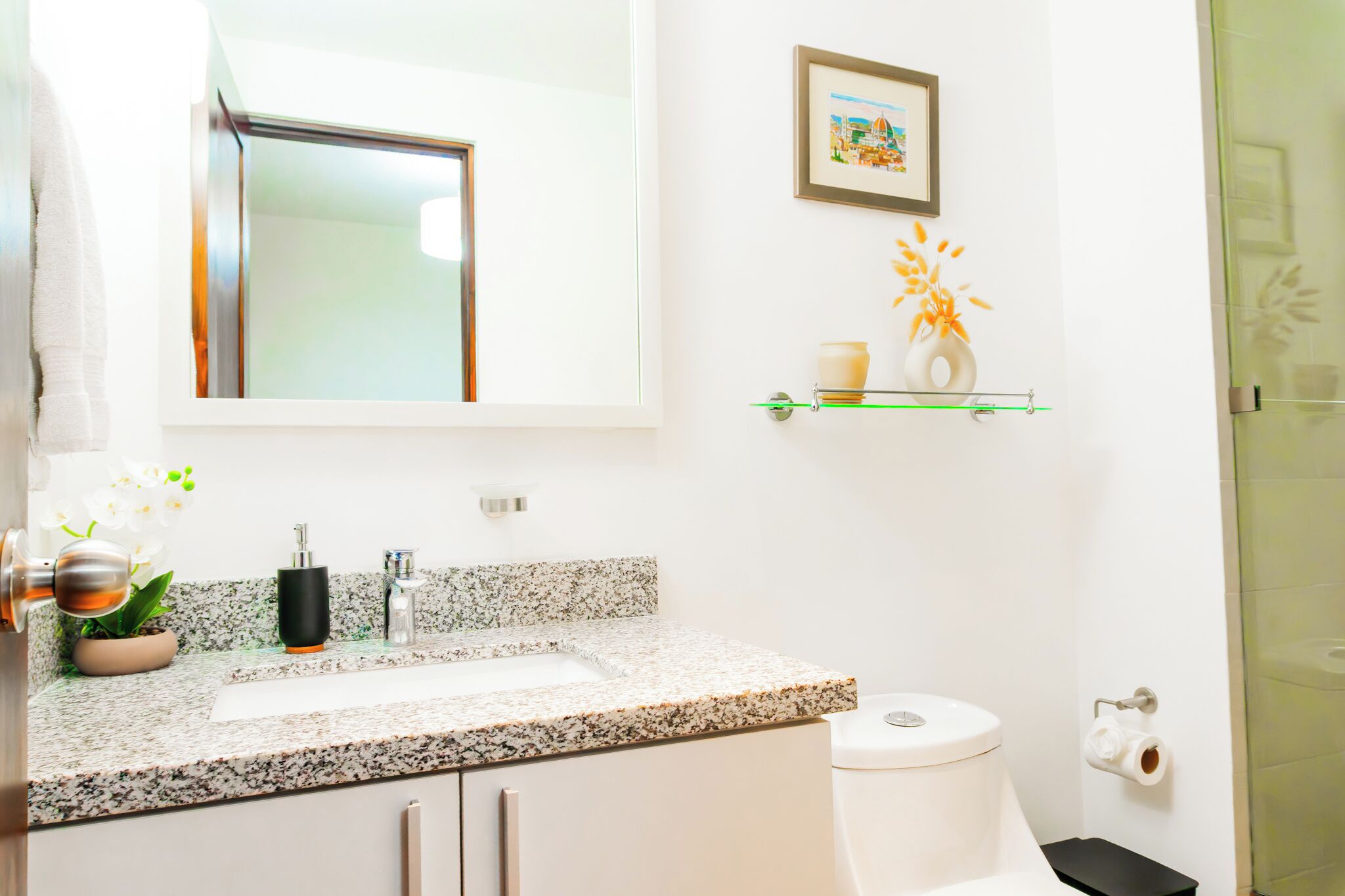 Un baño con encimera de granito, inodoro, lavabo, espejo, un cuadro en la pared y un pequeño estante con un florero y una vela.