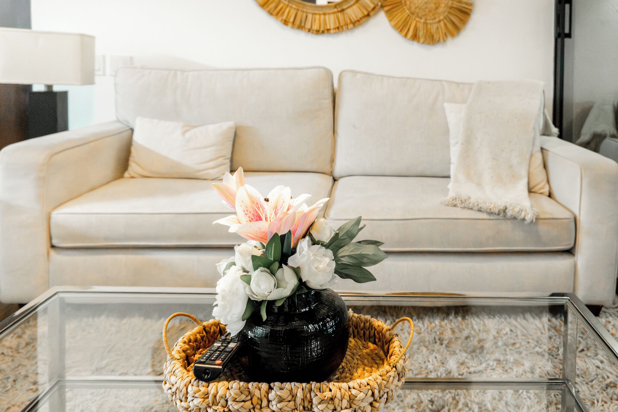 Una sala de estar con un sofá, una mesa de centro de vidrio, una cesta tejida y un florero con flores.