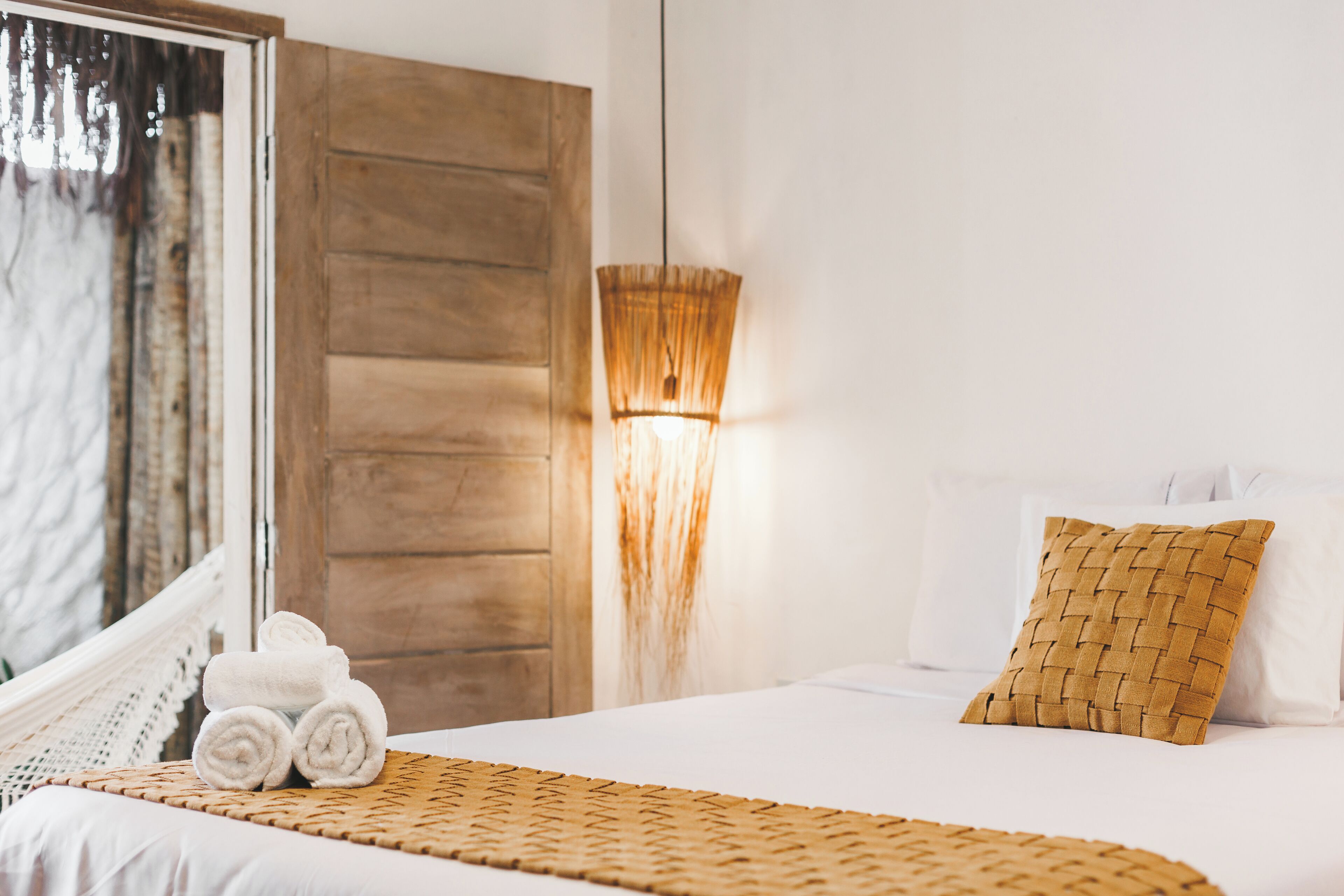 A neatly made bed with a woven blanket, a woven pillow, and rolled towels on a white bedspread.