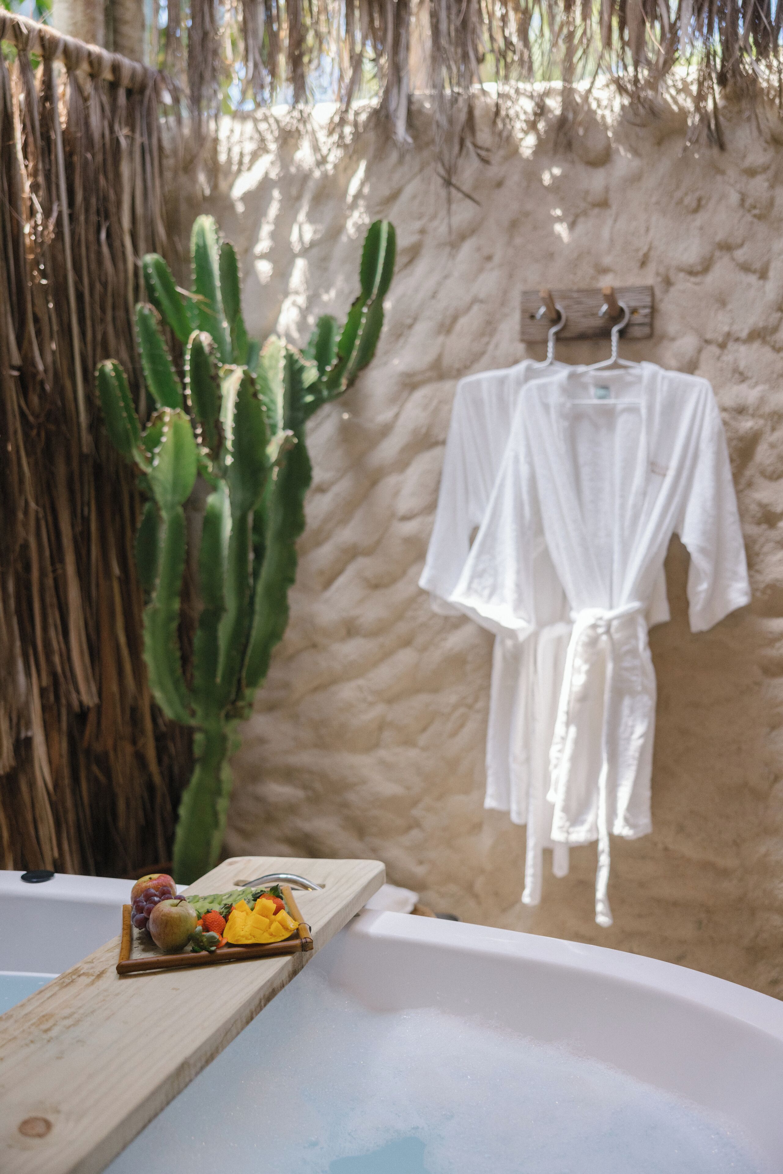 A bathtub with a wooden tray holding fruits and a white robe hanging on a wooden rack.