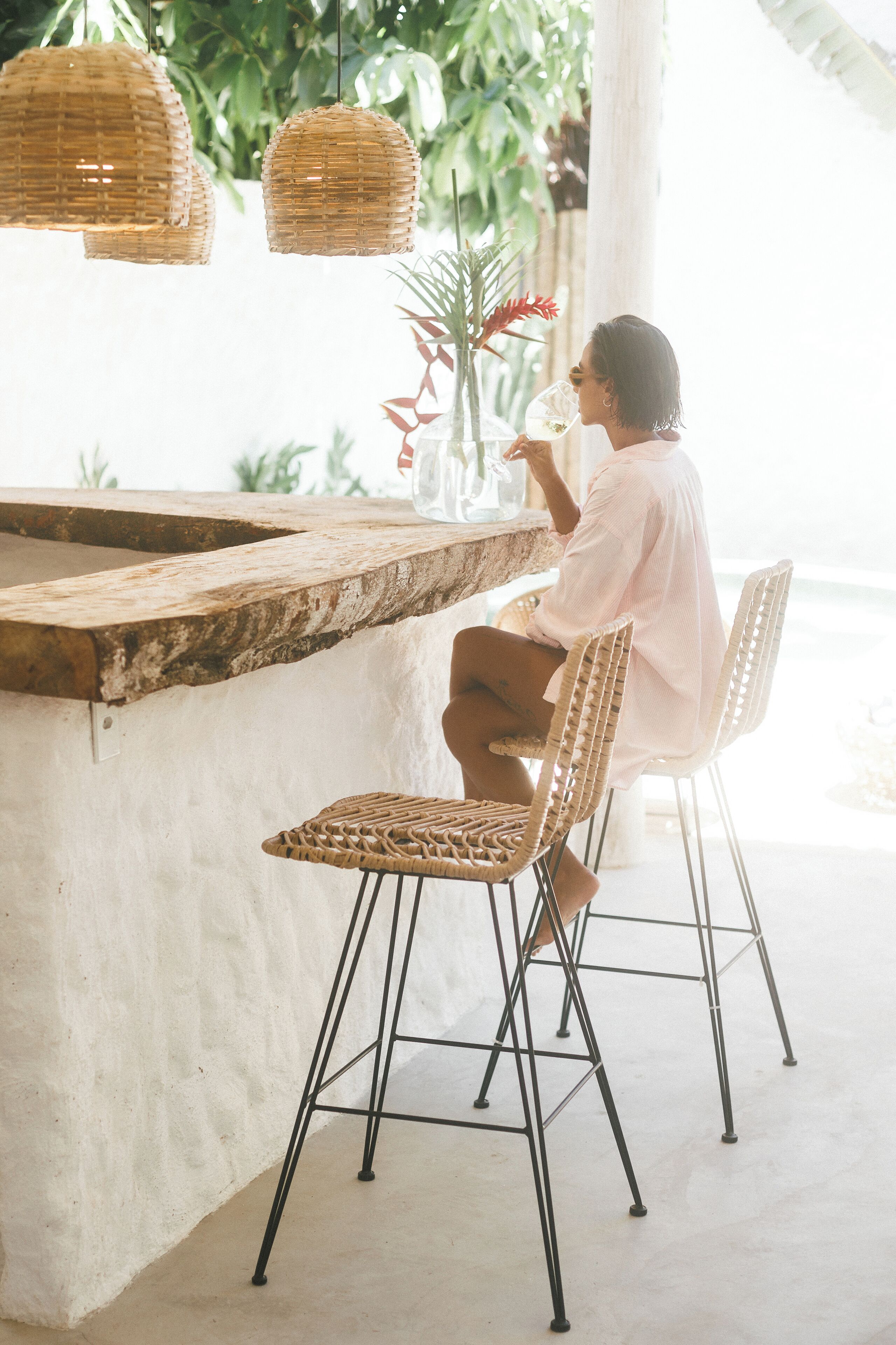 A person seated at a table with a woven chair, a wicker pendant light, and a white wall.