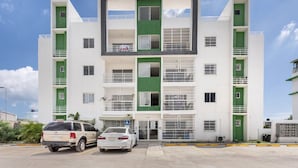 A modern multi-story building with a green and white color scheme, featuring balconies and a parking area with two cars.