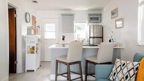 A compact kitchen with white cabinets, a stainless steel refrigerator, and a microwave. There are two wooden chairs at a small island, and a teal sofa with patterned cushions. - Charming 1-Bedroom Oasis in the Heart of Downtown (New Port Richey)