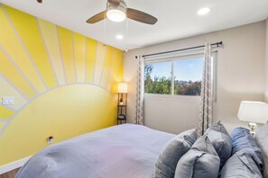 Ein Schlafzimmer mit einem Bett, einem Deckenventilator, einem Fenster mit Vorhängen und einer Wand mit Sonnenelement.