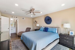 Ein Schlafzimmer mit Bett, Sessel, Nachttisch, Lampe, Deckenventilator und Wandbild.