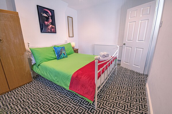 A bedroom with a patterned floor, a bed with green and red bedding, a peacock pillow, and a framed picture on the wall.