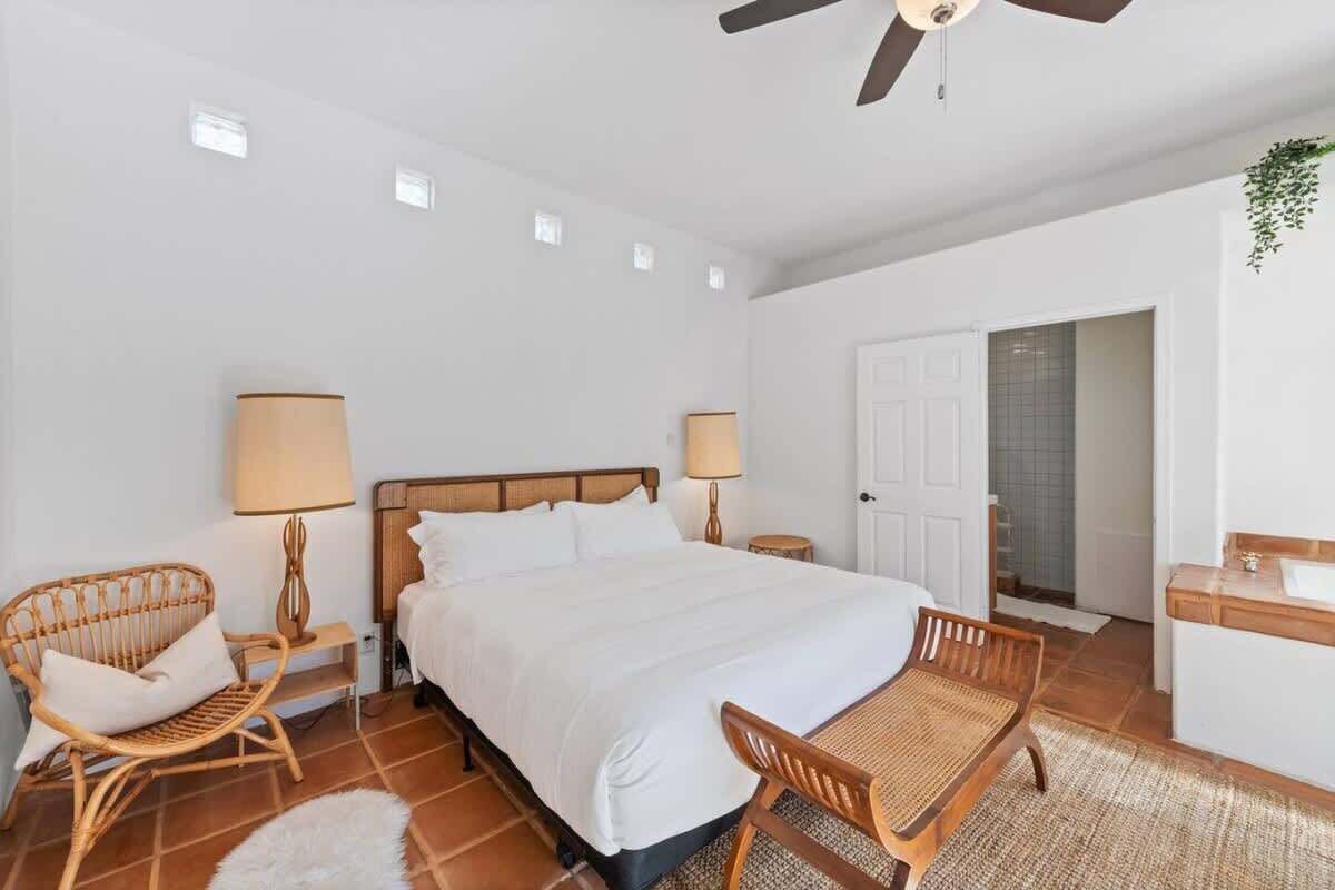 A bedroom with a large bed, two bedside tables with lamps, a wooden chair, a small table, and a door leading to another room.