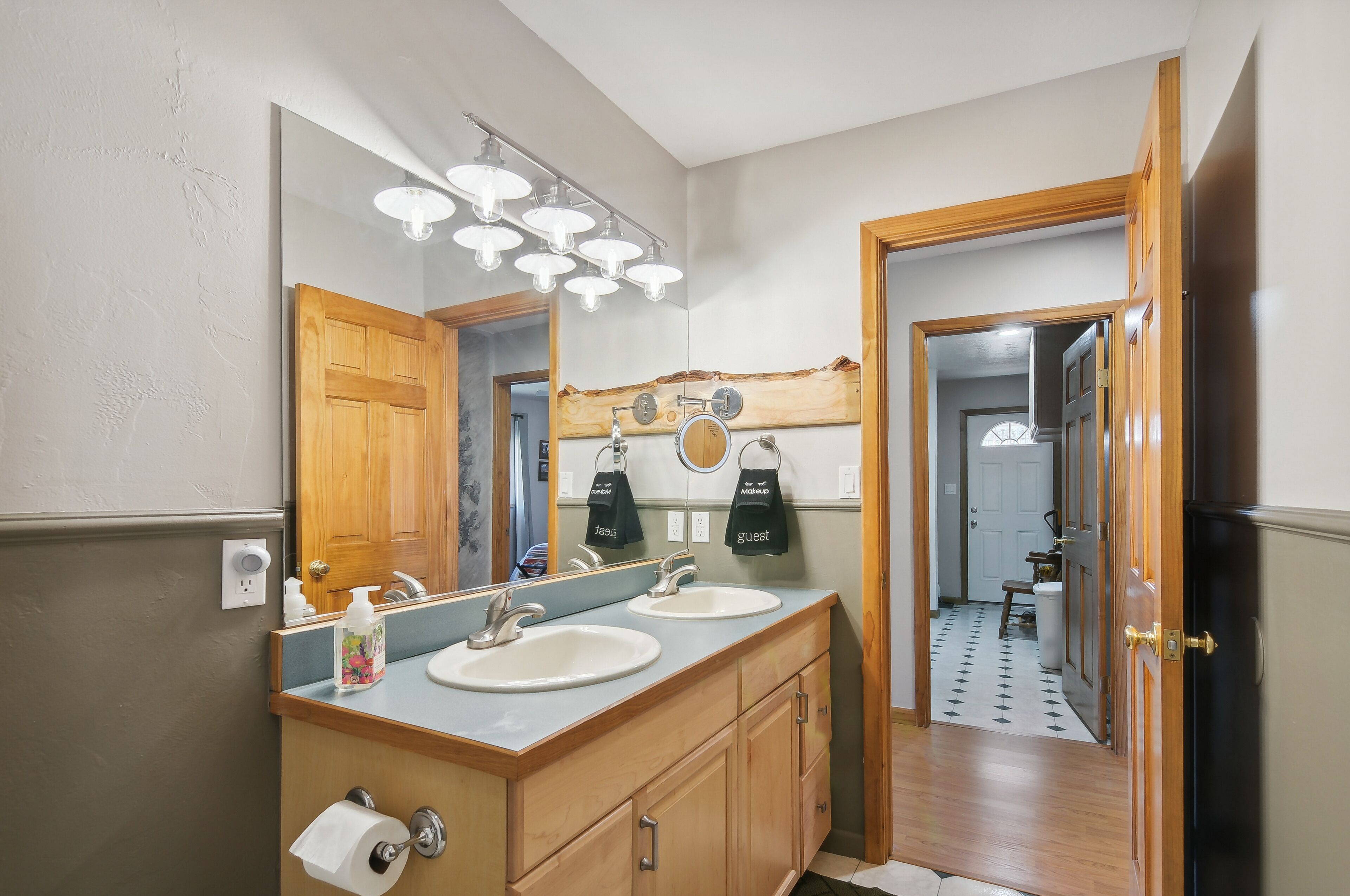 A bathroom with two sinks, a large mirror, and a wooden vanity. There are towels hanging on the wall and a toilet paper holder.