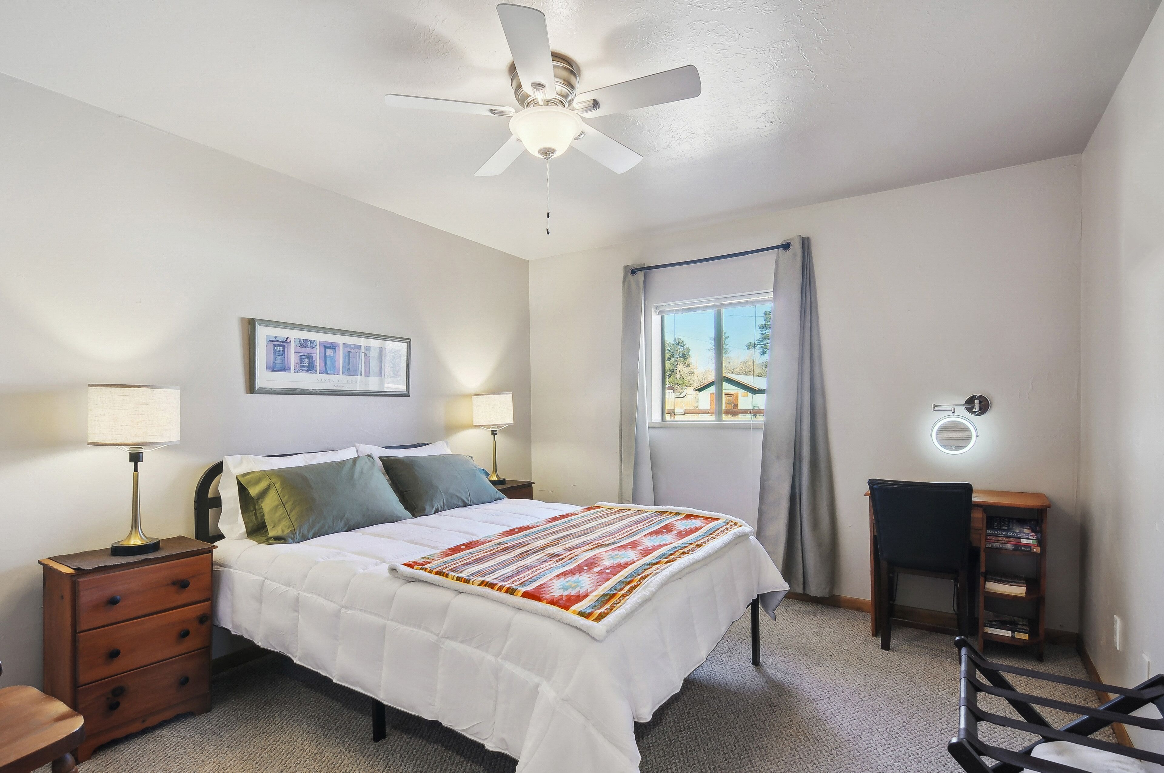 A bedroom with a bed, nightstands, a desk, a chair, a ceiling fan, a window with curtains, and a wall-mounted light.