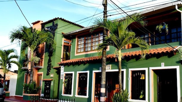 A green building with multiple windows and doors, a tiled roof, and a small fenced area with palm trees. - Apartahotel Tierras Amazonicas (Leticia)