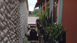 A narrow corridor with a textured wall on one side and a building with a red and green facade on the other. There are potted plants along the corridor and a metal railing on the right. - Apartahotel Tierras Amazonicas (Leticia)