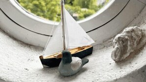 Um modelo de veleiro pequeno sobre uma superfície texturizada, uma estatueta de pássaro e um objeto semelhante a pedra, tudo dentro de uma moldura circular.