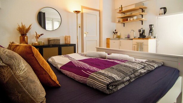 Quarto com cama, almofadas, mesa de cabeceira, espelho, guarda-roupa e cozinha equipada com prateleiras e eletrodomésticos.