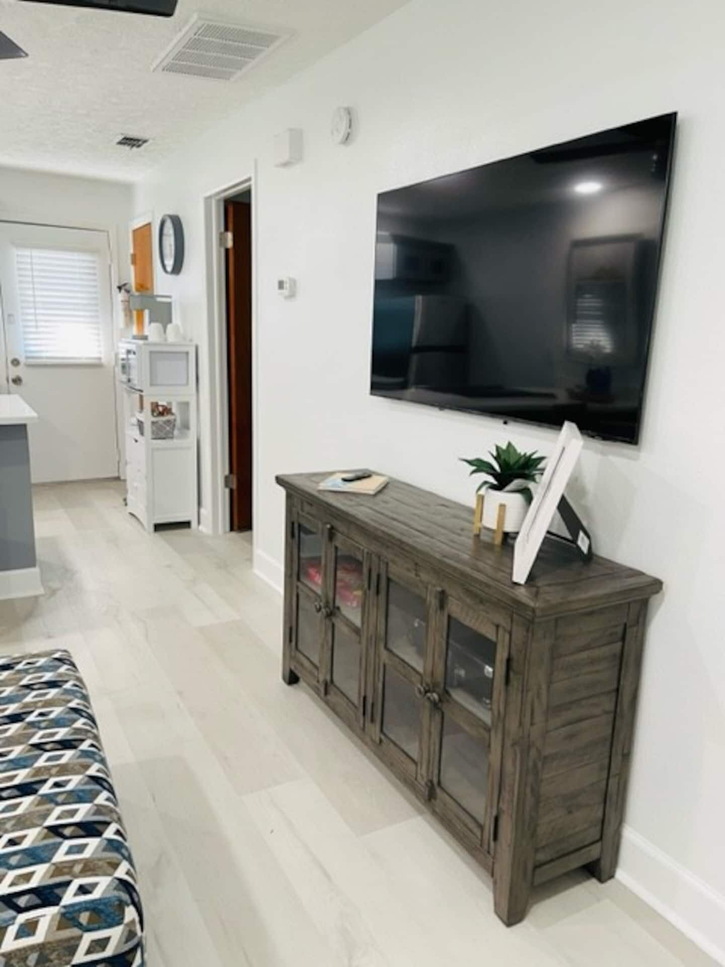 A hotel room with a large flat-screen TV mounted on the wall, a wooden cabinet with glass doors, a patterned bed, and a kitchenette area.