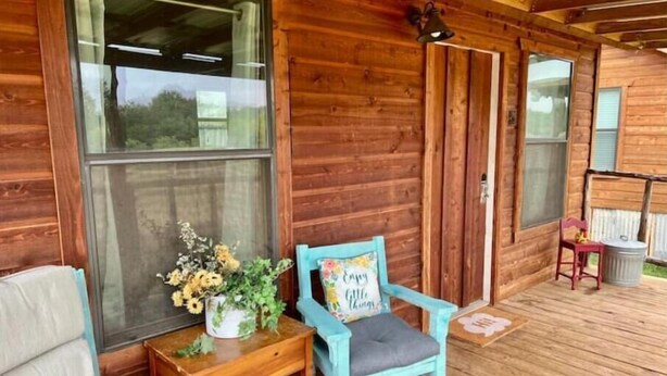 Une cabane en bois avec un porche, une chaise bleue avec un coussin, une petite table ornée d’un vase de fleurs et une fenêtre donnant sur un paysage verdoyant.