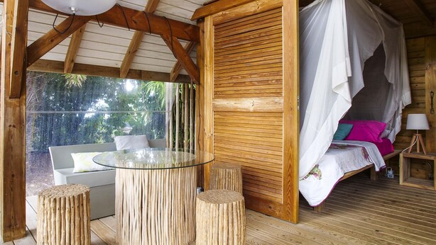 A wooden cabin with a glass-enclosed dining area, a bed with a canopy, and a small table with stools. - Villa Tiki a harmonious composition with a charming bungalow and private pool (Le Moule)