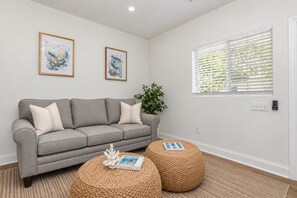 A living room with a grey sofa, two round woven ottomans, a framed abstract painting, a potted plant, and a window with blinds.