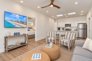 A modern living room with a flat-screen TV, a sofa, two striped armchairs, a coffee table, and a kitchen area with white cabinets and a refrigerator.