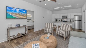 Ein modernes Wohnzimmer mit großem Fernseher, Deckenventilator und Küchenbereich mit weißen Schränken und Edelstahlgeräten.