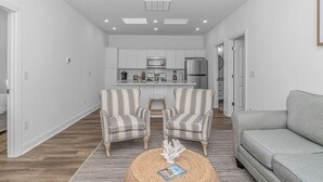 Eine moderne Wohnung mit einem zentralen Wohnbereich, ausgestattet mit zwei gestreiften Sesseln, einem grauen Sofa und einem gewebten Couchtisch. Im Hintergrund ist eine Küche mit weißen Schränken und Edelstahlgeräten zu sehen.