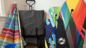 Une pièce avec un plancher en bois, une couverture rayée aux couleurs vives, un seau bleu, une pelle bleue, une valise grise et un sac à dos coloré.