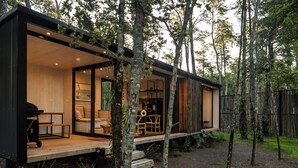 Una cabaña moderna con terraza de madera, asientos integrados y un amplio ventanal con vistas al bosque.