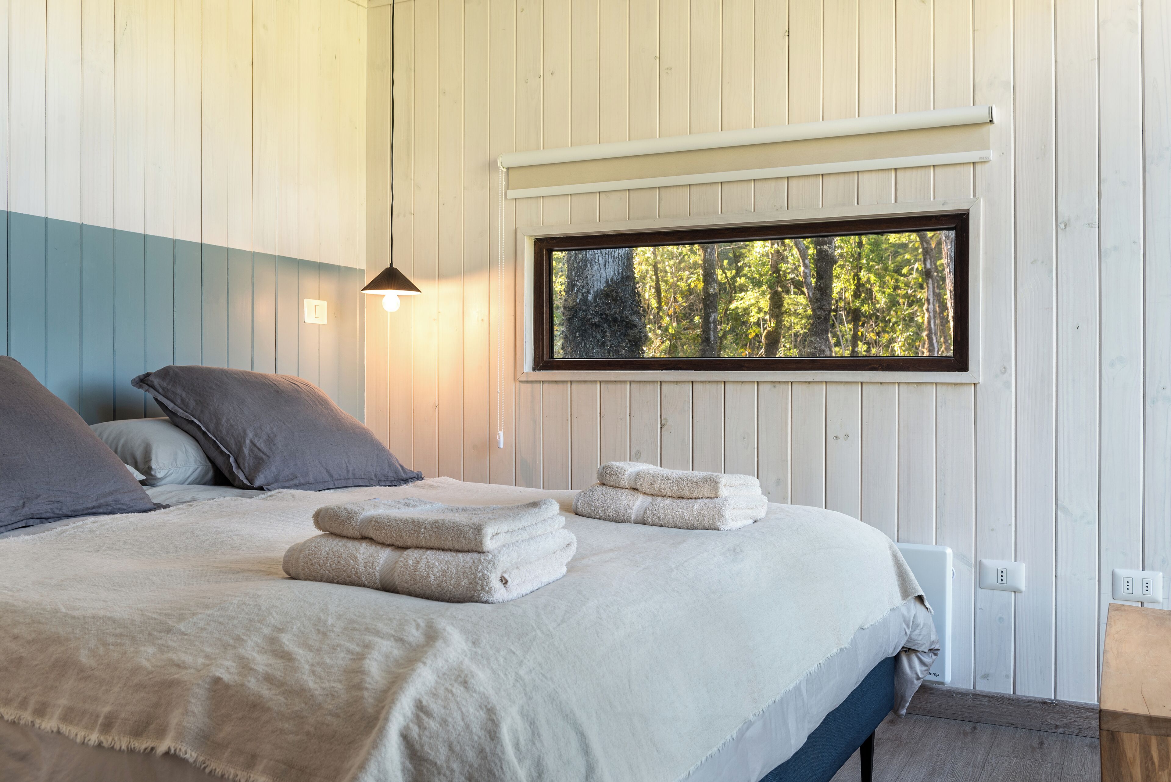 A bedroom with a bed, pillows, a window with a view of trees, and a bedside lamp.
