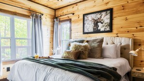 A wooden cabin bedroom with a bed, pillows, a blanket, a framed picture on the wall, and a window with curtains. - 3 King Beds 3.5 Bath Cabin-Hot Tub-Pool Table, Great Smoky Mountains (Sevierville)